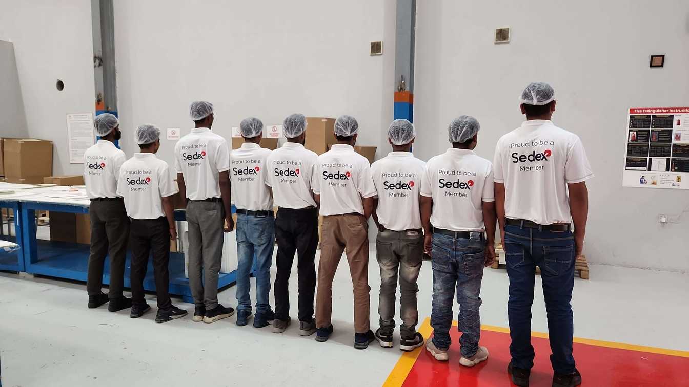 A group of workers in a warehouse stand with their backs to the camera, wearing white shirts that say "Proud to be a Sedex Member" and hair nets. They are positioned in front of a workbench and boxes. This image represents Sedex certification in Pakistan, highlighting compliance with ethical and responsible business practices.